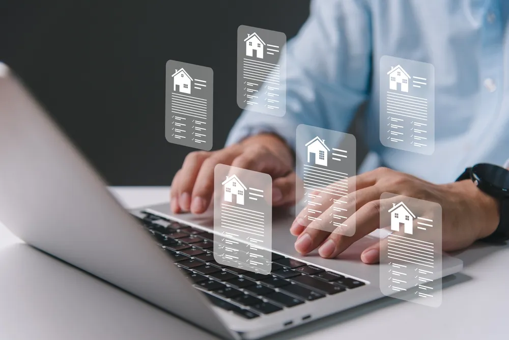Shoulder-down image of a man sitting at a laptop typing. Hovering above the man's hands on the laptop are six lightly transparent-white home data sheets. The data sheets each contain the outline of a house, lines that indicate lists, and check mark next to some of the line items.